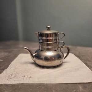 May include: A silver-colored coffee pot with a matching lid and a small cup stacked on top. The pot sits on a white embroidered cloth. The background is a solid gray color.