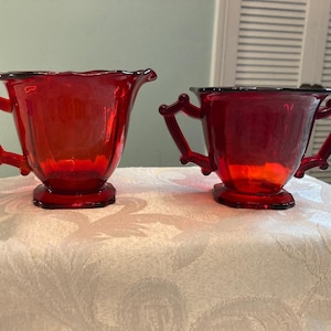 May include: A pair of ruby red glass vessels, consisting of a creamer with a single handle and a sugar bowl with two handles. Both pieces feature a scalloped rim, a faceted body, and a square base, showcasing a deep, translucent red color.