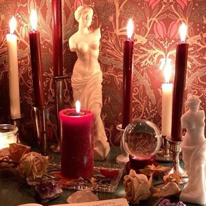 K&ouml;nnte beinhalten: Eine rote Kerze brennt hell auf einem Tisch mit einer Kristallkugel, zwei wei&szlig;en Kerzen und zwei Statuen. Der Tisch ist mit einer rot-goldenen gemusterten Tischdecke bedeckt. Auf dem Tisch liegt ein Tagebuch mit handschriftlichem Text.