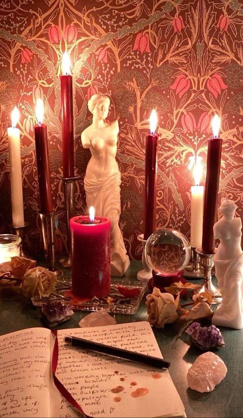 K&ouml;nnte beinhalten: Eine rote Kerze brennt hell auf einem Tisch mit einer Kristallkugel, zwei wei&szlig;en Kerzen und zwei Statuen. Der Tisch ist mit einer rot-goldenen gemusterten Tischdecke bedeckt. Auf dem Tisch liegt ein Tagebuch mit handschriftlichem Text.