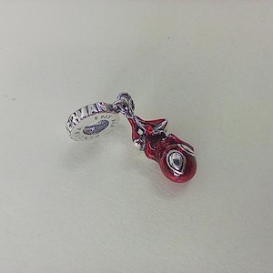 May include: A silver charm with a red, glossy design. The charm features a round, red element with a silver eye-like detail. The charm is attached to a silver ring with the word "ERMAN" visible. It is likely a jewelry accessory.