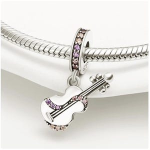 Puede incluir: Un charm de plata con un colgante de viol&iacute;n blanco adornado con piedras preciosas rosas y moradas. El charm cuelga de una pulsera de plata. El viol&iacute;n tiene cuerdas y un arco detallados, mostrando un tema musical.