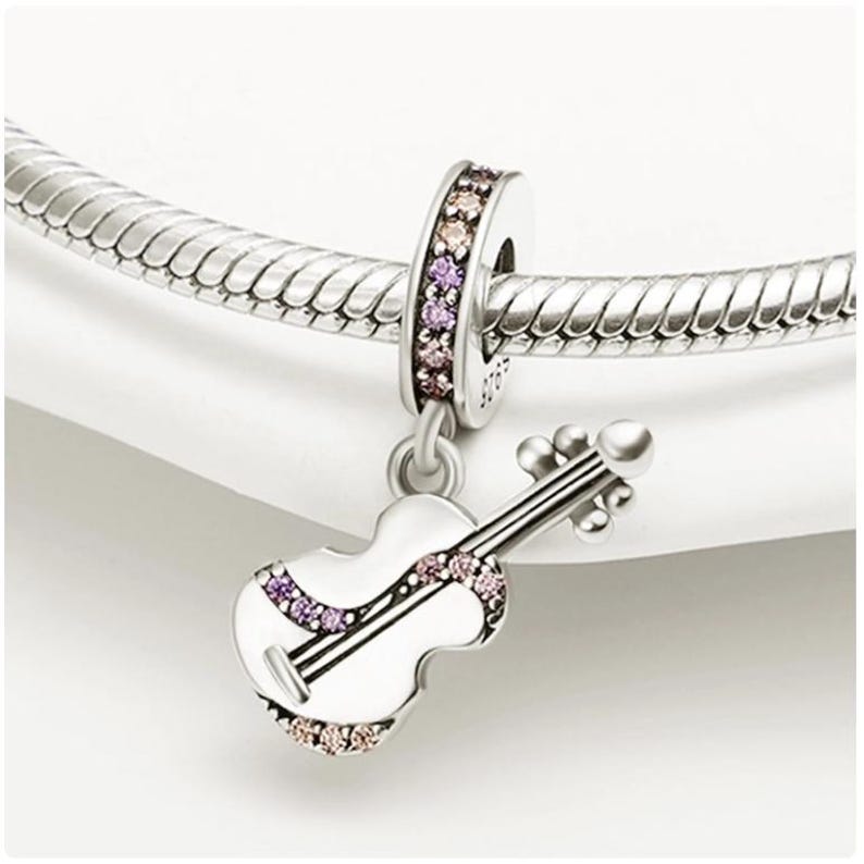 Puede incluir: Un charm de plata con un colgante de viol&iacute;n blanco adornado con piedras preciosas rosas y moradas. El charm cuelga de una pulsera de plata. El viol&iacute;n tiene cuerdas y un arco detallados, mostrando un tema musical.