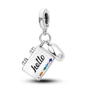 Puede incluir: Un charm plateado con diseño de cuaderno, la palabra "hello" en cursiva y una fila de gemas coloridas. Un pequeño charm redondeado con una gema transparente está adjunto. El charm está suspendido de un lazo plateado.