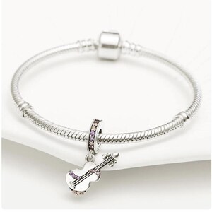 Puede incluir: Una pulsera de plata con un charm de viol&iacute;n. El charm de viol&iacute;n es blanco con detalles morados y transparentes. El charm tambi&eacute;n tiene una banda plateada con detalles morados y transparentes. La pulsera est&aacute; sobre una superficie blanca.
