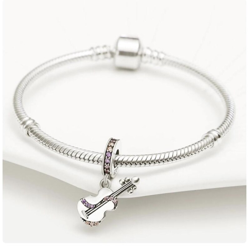 Puede incluir: Una pulsera de plata con un charm de viol&iacute;n. El charm de viol&iacute;n es blanco con detalles morados y transparentes. El charm tambi&eacute;n tiene una banda plateada con detalles morados y transparentes. La pulsera est&aacute; sobre una superficie blanca.