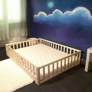 May include: A light-colored wooden toddler bed with a slatted frame and a white mattress. The bed is placed on a dark wood floor, with a white rug nearby. The wall behind the bed is painted with a night sky scene.