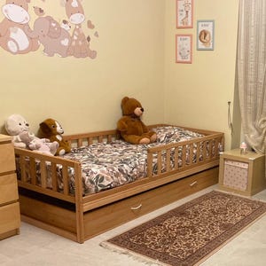 Peut inclure: Lit pour enfant en bois clair avec une couette à motifs floraux et un tiroir coulissant. Le lit est entouré d'animaux en peluche et repose sur un tapis à motifs. La pièce a un mur jaune clair avec des décorations murales sur le thème des animaux.