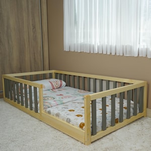 May include: A wooden toddler bed with gray and natural wood slats. The bed is made of wood and has a mattress with a colorful patterned sheet.