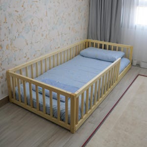 Könnte beinhalten: Ein Kinderbett aus Holz mit einer hellblauen Matratze und einem weißen Kissen. Das Bett hat an allen Seiten ein Sicherheitsgitter.