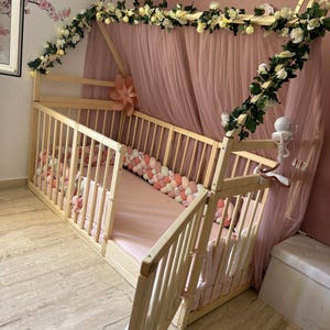 May include: A light wood baby crib with a canopy draped in pink tulle. The crib is decorated with white flowers and greenery. A pink and white braided bumper lines the inside of the crib.