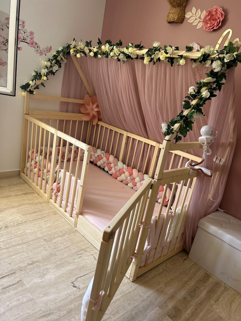 May include: A light wood baby crib with a canopy draped in pink tulle. The crib is decorated with white flowers and greenery. A pink and white braided bumper lines the inside of the crib.
