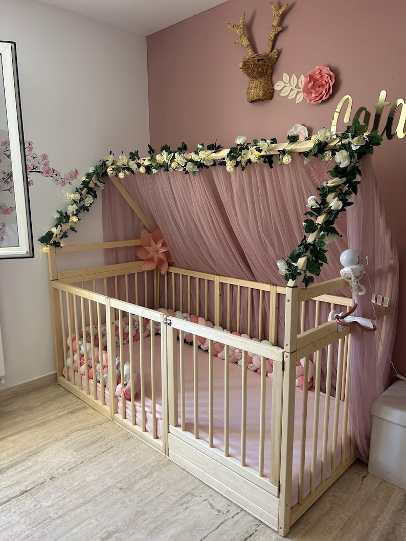 May include: A light wood baby bed with a canopy draped in pink tulle. The bed is decorated with a garland of white flowers and greenery. The bed is in a room with pink walls and a decorative deer head.