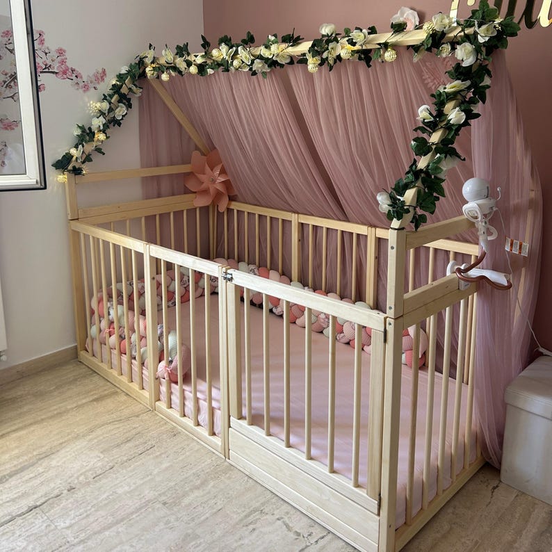 May include: A light wood baby crib with a canopy draped in pink tulle. The crib is decorated with white flowers and greenery. The crib is filled with pink pillows and a pink mattress.