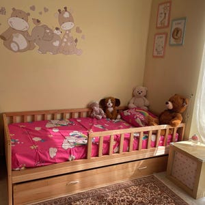 May include: A wooden toddler bed with a pink patterned duvet cover and a pull-out drawer. The bed is surrounded by soft toys, including a teddy bear, a dog, and a white bear. The wall features cartoon animal decals.
