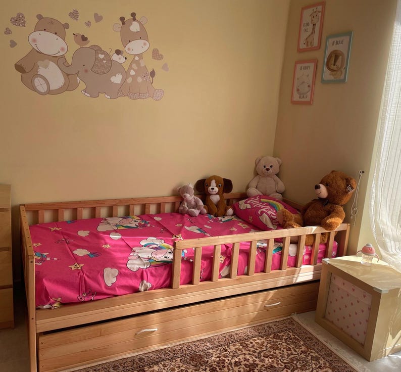 May include: A wooden toddler bed with a pink patterned duvet cover and a pull-out drawer. The bed is surrounded by soft toys, including a teddy bear, a dog, and a white bear. The wall features cartoon animal decals.