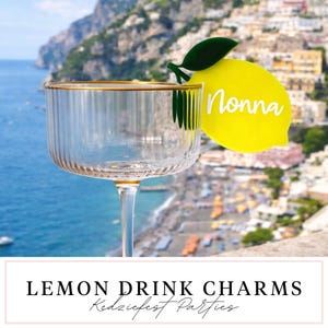 Puede incluir: Un vaso transparente con borde dorado y un adorno para bebidas de limón amarillo que dice "Nonna". El fondo muestra un pueblo costero con un mar azul. El texto "Lemon Drink Charms" está en la parte inferior de la imagen.