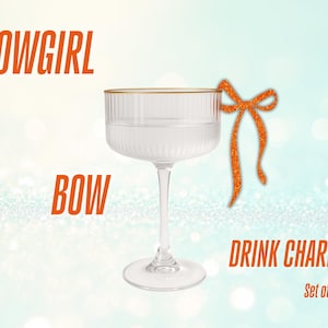 Puede incluir: Una copa de cristal transparente con borde dorado y un lazo decorativo de purpurina naranja. Las palabras "SHOWGIRL BOW DRINK CHARMS" están en texto naranja. La imagen está sobre un fondo azul claro.