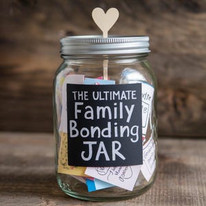 May include: A clear glass jar with a silver lid, filled with colorful notes. A wooden heart is attached to a stick on top. The jar has a black label that reads "THE ULTIMATE Family Bonding JAR".