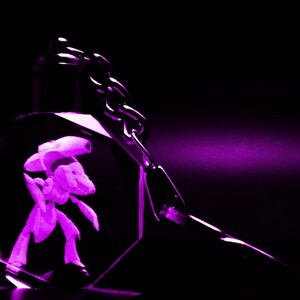 May include: A purple crystal keychain featuring a 3D image of a character. The keychain has a silver chain and a metallic clasp. The image is illuminated with a purple light against a black background.