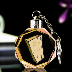 May include: A novelty keychain with a cartoon character, a smiling piece of wood. The keychain has a faceted, jewel-like body with a silver-coloured top and chain. The background is dark, highlighting the colourful keychain.