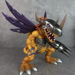 Puede incluir: Figura detallada de un personaje de Digimon en una pose dinámica. La figura es principalmente amarilla con detalles azules, armadura plateada metálica y alas moradas. Tiene garras afiladas, cuernos y una expresión amenazante.