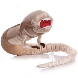 May include: A plush toy of a creature from the film Alien. The toy is beige and has a large, open mouth with red lips and white teeth. The creature has a long, segmented body and a tail.
