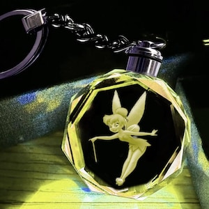 May include: A faceted, clear crystal keychain featuring a laser-etched, glowing yellow illustration of a winged fairy character holding a small wand. The crystal is attached to a silver-toned metal chain and a circular split ring for keys.