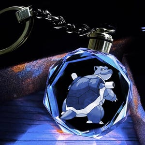 May include: A clear, multifaceted crystal keychain featuring a laser-engraved image of the blue turtle-like creature Blastoise. The pendant is attached to a silver-toned metal chain and a circular split ring, showcasing a detailed 3D character design.