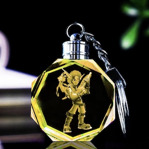 May include: A gold-colored crystal keychain featuring a cartoon character holding a weapon. The character is a green-skinned elf with large ears and a red hat. The keychain is shaped like a hexagon and has a silver metal ring.