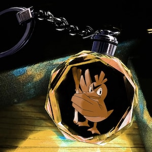 May include: A faceted, circular crystal keyring featuring a brown, cartoon-style bird character holding a long, light-coloured stalk. The crystal has a metallic silver top attachment connected to a silver-toned metal chain and a circular key ring.