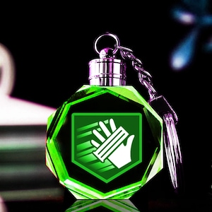 Peut inclure: Un porte-clés en cristal vert facetté avec une chaîne et un fermoir argentés. Le porte-clés présente un emblème vert brillant avec un motif de main et de lignes. Le fond est sombre, avec des touches de violet et de bleu.