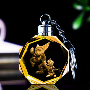 Op de afbeelding: Een gouden kristallen sleutelhanger met een 3D-afbeelding van Ratchet en Clank uit de videogame-serie. De sleutelhanger is zeshoekig en heeft een zilveren metalen ring.