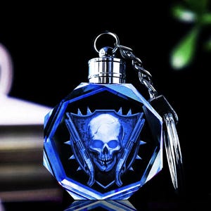 May include: A blue crystal keychain with a skull and crossbones design. The skull is white and the crossbones are silver. The keychain is attached to a silver ring.
