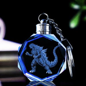 May include: A blue crystal keychain with a silver metal ring and a 3D laser engraved image of Godzilla. The crystal is shaped like an octagon.