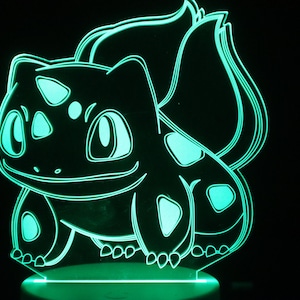 May include: A glowing green Bulbasaur LED lamp. The lamp features a detailed outline of the Pokémon character, with green accents on a black background. The lamp is designed to sit on a flat surface and emits a soft, ambient light.