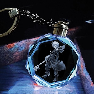 May include: A blue crystal keychain with a silver key chain and a 3D image of a character holding a key. The character is wearing a black coat and has white hair.