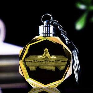 Peut inclure: Un porte-clés en cristal jaune avec une chaîne et un porte-clés argentés. À l'intérieur du cristal se trouve un cercueil miniature avec un ours en peluche et des points d'interrogation. Le porte-clés est octogonal et a un dessus argenté.