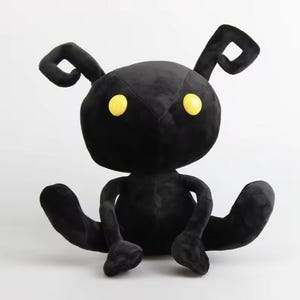 May include: Black plush toy shaped like a creature with large yellow eyes and two small ears. The toy is sitting with its legs crossed.