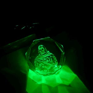 May include: A green crystal paperweight with a 3D etched image of a skeleton wearing a hooded cloak and holding a weapon.