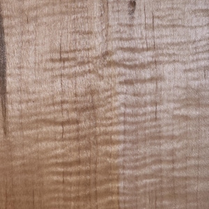 May include: Close-up of a piece of wood with a wavy grain pattern. The wood has a light brown color with darker streaks and knots. The grain creates a textured appearance.
