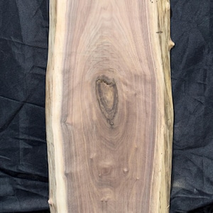 May include: A rectangular, unfinished wooden slab with a natural edge. The wood grain features a mix of light and dark brown tones, with a prominent knot in the center. The slab has a rough, natural edge on one side and a smooth, cut edge on the other.