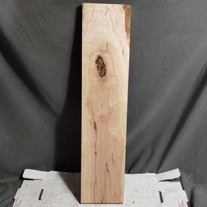 May include: A rectangular, unfinished wooden board with a light tan color and a prominent knot. The wood grain is visible, with darker streaks and patterns. The board is likely for woodworking or craft projects.