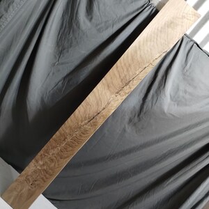 May include: A long, rectangular piece of light-colored wood with a natural grain pattern and a dark, linear crack running down the center. The wood has a smooth, finished appearance, and the grain creates a swirling, textured effect.