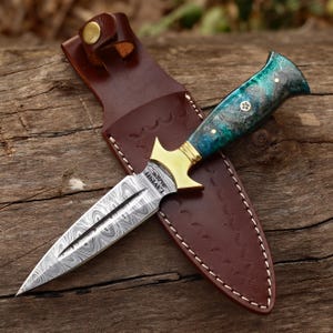 May include: A Damascus steel dagger with a patterned blade and a handle with blue and green accents. The dagger has a gold-colored guard and is in a brown leather sheath. The sheath has white stitching and a leather strap with a gold button.