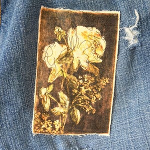 May include: Close-up of blue denim shorts with distressed details. A rectangular patch featuring a floral design with yellow and brown tones is sewn onto the shorts. The floral design includes roses and other flowers.