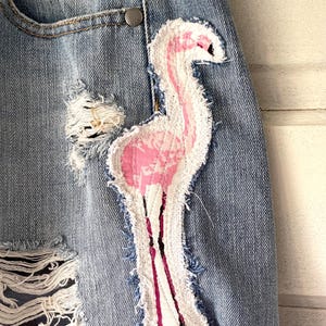 May include: Close-up of denim shorts with a distressed design. A pink and white flamingo patch is sewn onto the shorts. The shorts have a rolled hem and a button closure.