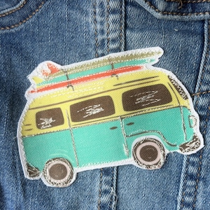May include: A fabric patch shaped like a vintage camper van with a yellow upper body and a teal lower body. The van features brown windows, dark wheels, and a roof rack carrying two surfboards in red and green. The patch has a white border.
