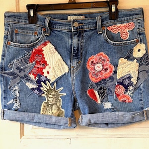 May include: Blue denim shorts with decorative patches. Patches include a star, floral designs, and a Statue of Liberty graphic. The shorts have rolled-up cuffs and are hanging on a black hanger.