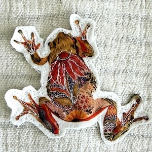 May include: A colorful fabric frog applique with a white border. The frog features a patterned design in shades of brown, red, and orange. The applique is sewn onto a textured, light gray fabric background.